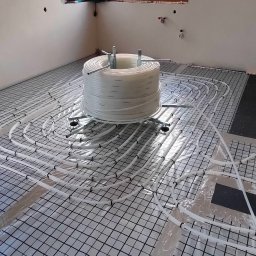 YOUVENT Michał Graczek - Rozłożona instalacja ogrzewania podłogowego na folii z rastrem, z rolką rur na stojaku w tle. Widoczne przygotowania do wylewki.