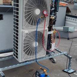 YOUVENT Michał Graczek - Dwuczęściowa jednostka zewnętrzna klimatyzacji na dachu, podłączona do pompy próżniowej. W tle wieża kościoła i zabudowa miejska.