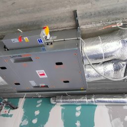 YOUVENT Michał Graczek - Szara centrala wentylacyjna Clima Gold zamontowana pod sufitem, z podłączonymi przewodami wentylacyjnymi w srebrnej izolacji. Widoczna drabina i ślady prac wykończeniowych.