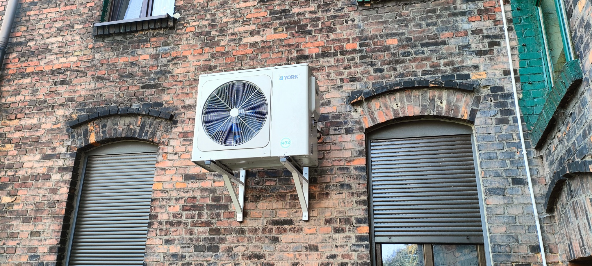 Biała jednostka zewnętrzna klimatyzacji YORK zamontowana na wspornikach na ceglanej ścianie budynku z oknami z żaluzjami. Widoczne ślady zabrudzeń na cegłach.