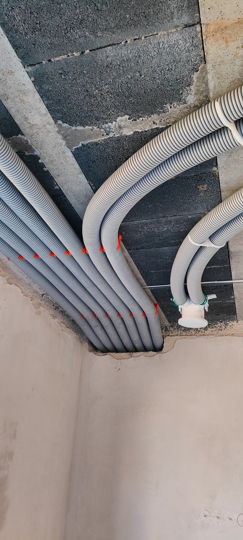 Szereg szarych, karbowanych rur wentylacyjnych przymocowanych do sufitu z betonu komórkowego za pomocą pomarańczowych uchwytów. Widoczny fragment ściany.