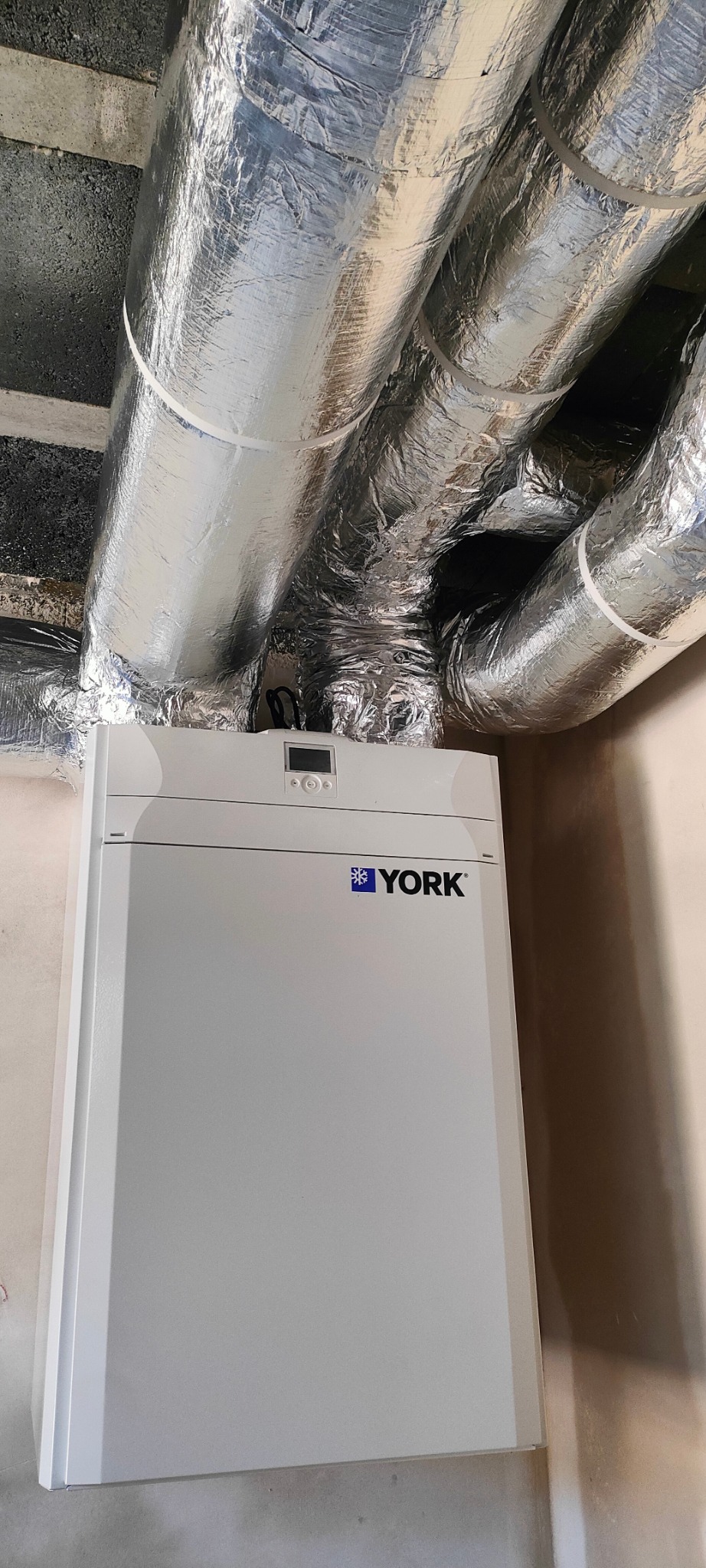 Biała jednostka klimatyzacyjna York z ekranem i rurami w izolacji aluminiowej pod sufitem. Widok z dołu, instalacja w pomieszczeniu.