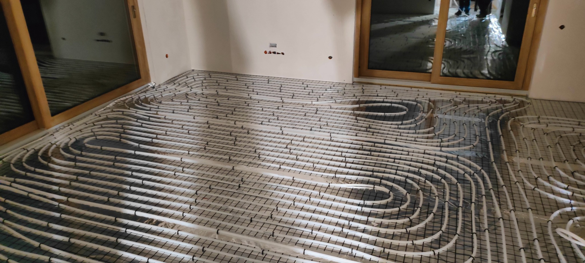 Białe rury ogrzewania podłogowego ułożone na czarnej siatce w jasnym pomieszczeniu z drewnianymi oknami. Widok z góry na instalację przed wylaniem posadzki.