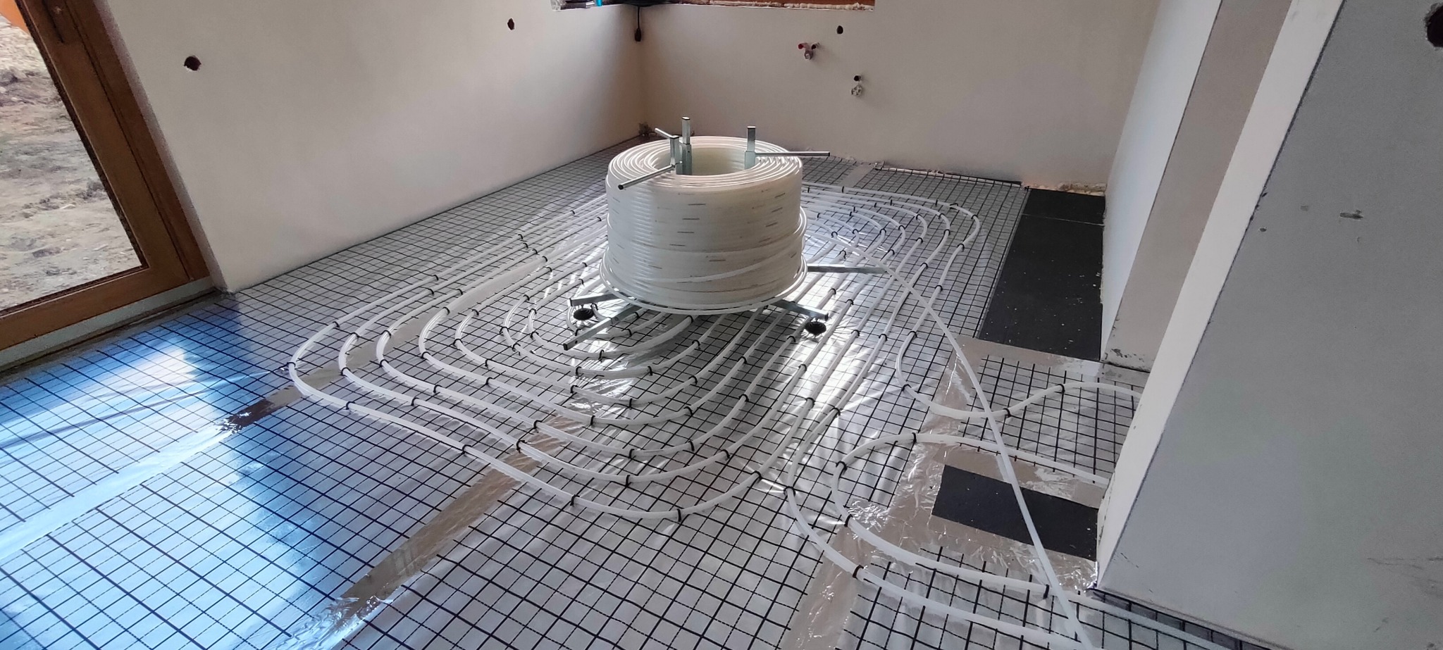 Rozłożona instalacja ogrzewania podłogowego na folii z rastrem, z rolką rur na stojaku w tle. Widoczne przygotowania do wylewki.