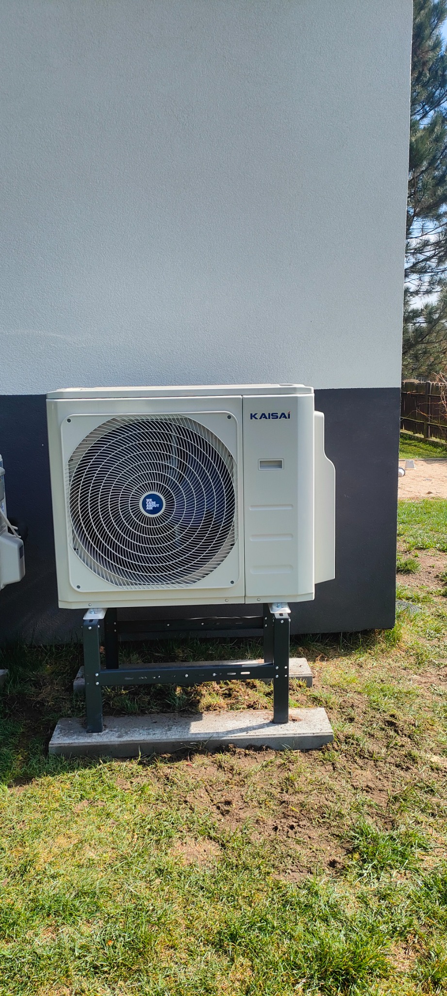 Biała jednostka zewnętrzna klimatyzacji Kaisai, zamontowana na metalowym stelażu na trawniku przy budynku z szarą elewacją. Widoczny fragment drugiej jednostki.