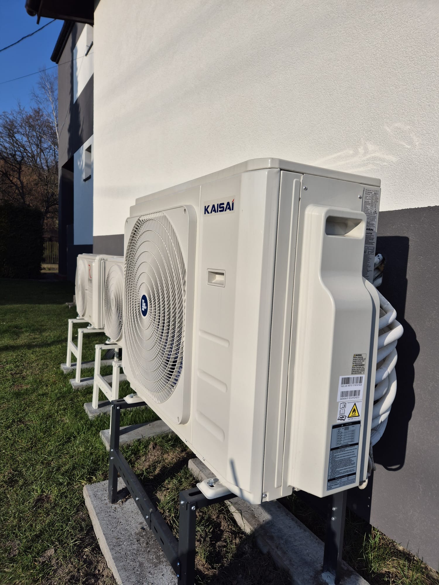 Białe jednostki zewnętrzne klimatyzacji Kaisai, zamontowane na metalowych wspornikach na trawniku przed budynkiem z szarą elewacją. Widok z boku, w tle drzewa.