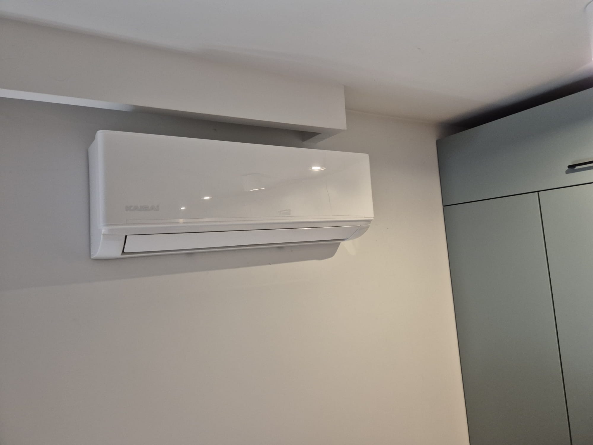 Biały klimatyzator marki Kaisai zamontowany na jasnej ścianie w nowoczesnym wnętrzu. Widoczny fragment zabudowy meblowej w odcieniach szarości.