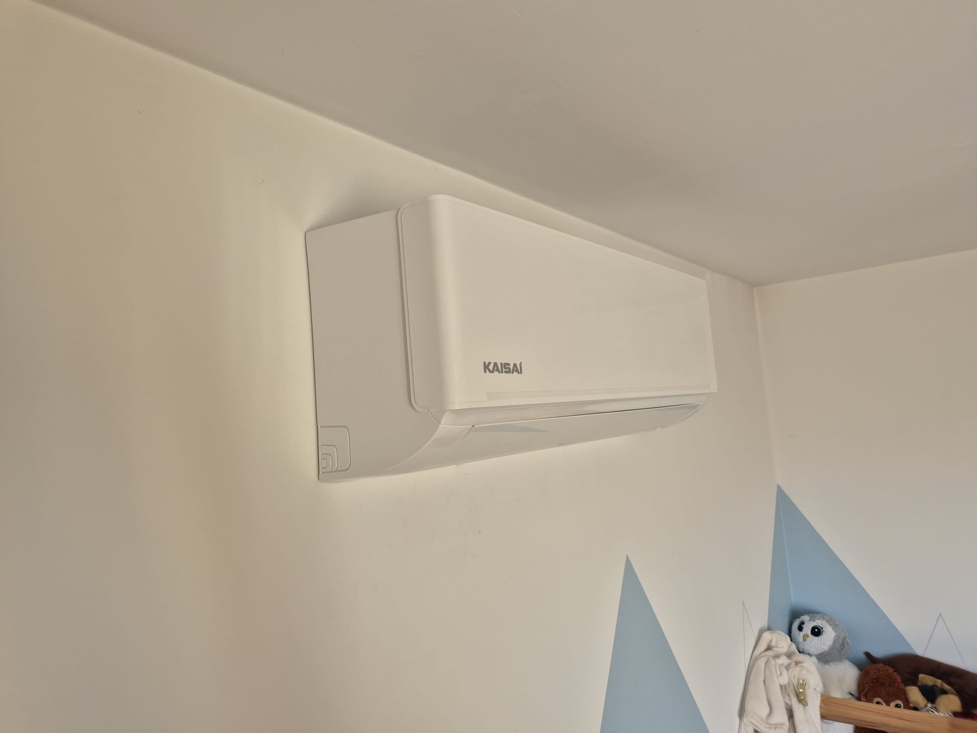 Biały klimatyzator Kaisai zamontowany na ścianie w pokoju dziecięcym z dekoracyjnymi, niebieskimi trójkątami i półką z pluszakami. Widok z dołu.