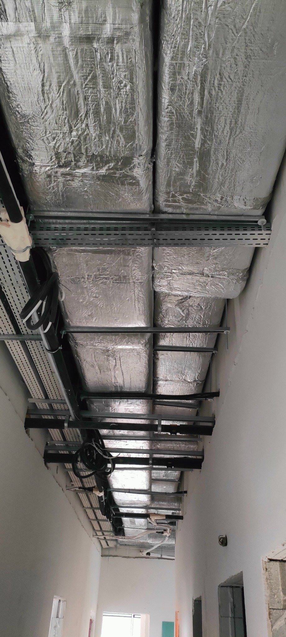 Widok z dołu na zamontowane, izolowane kanały wentylacyjne pod sufitem, z widocznymi elementami mocującymi i przewodami. Instalacja w surowym wnętrzu budynku.