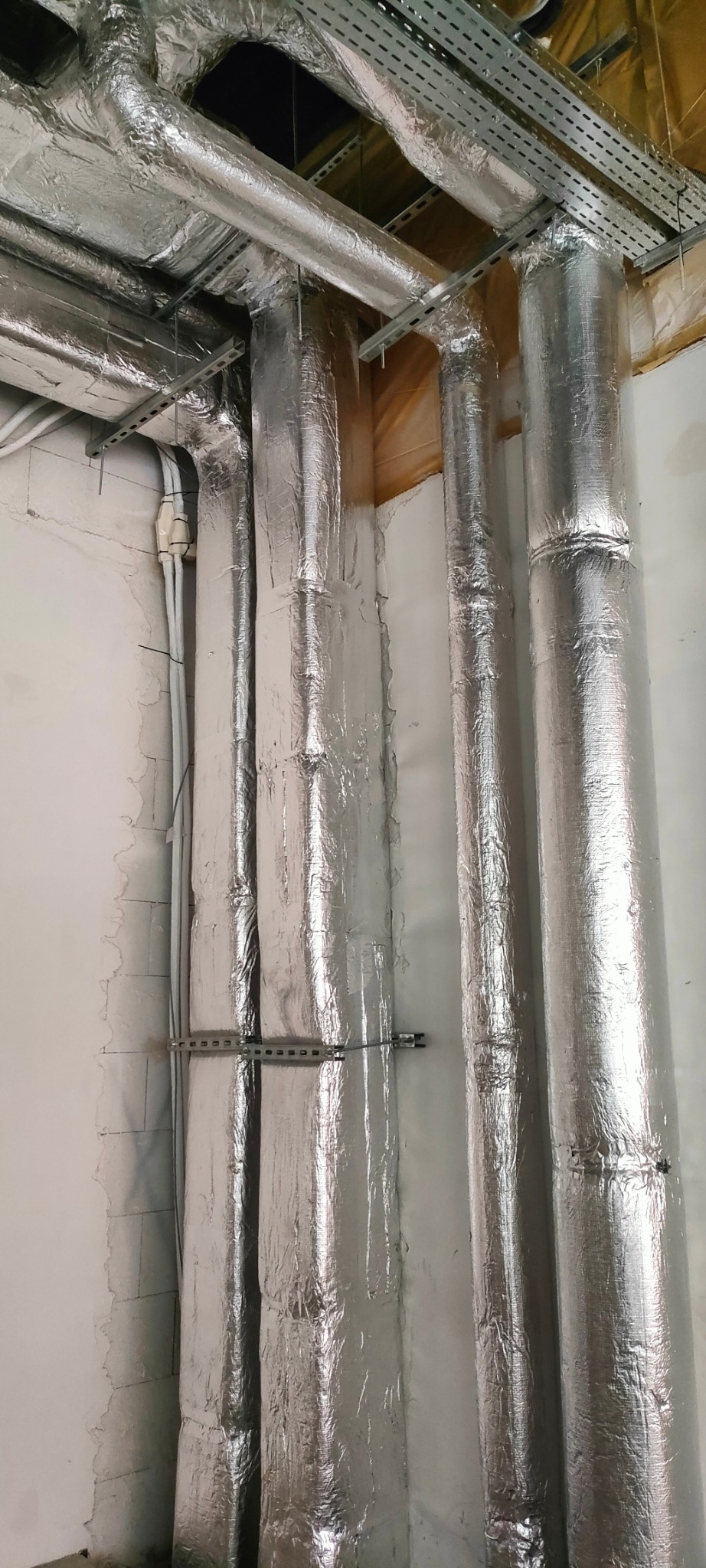Izolowane, srebrne rury wentylacyjne zamontowane przy ścianie i pod sufitem. Widoczne elementy mocujące i fragmenty instalacji elektrycznej.