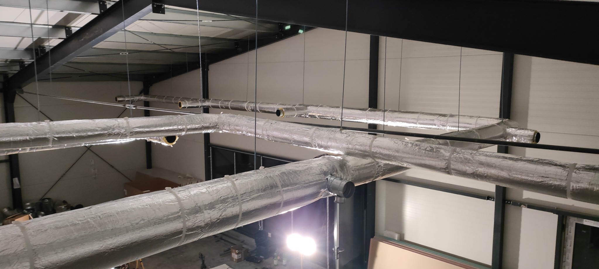 Izolowane rury wentylacyjne pod sufitem hali, widoczne elementy mocujące. Instalacja w srebrnej izolacji, czarne rury. Widok z góry na system wentylacyjny.