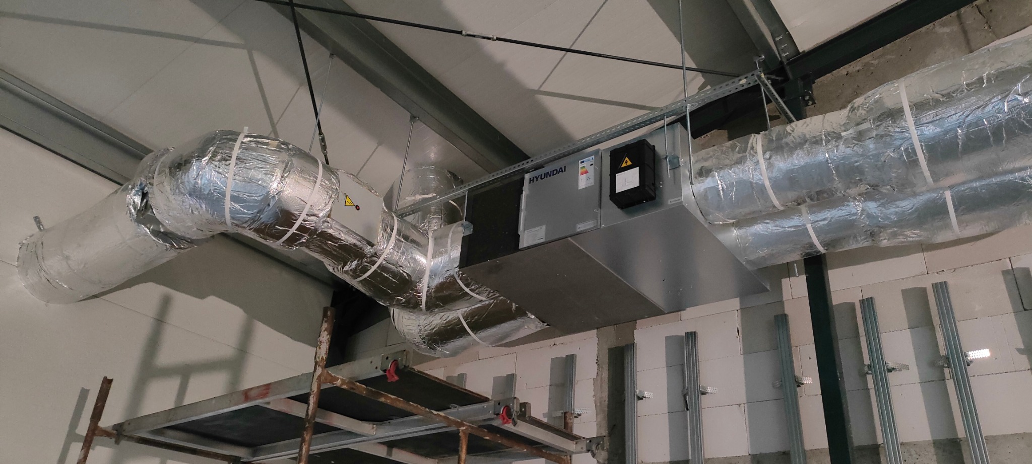 Zamontowana centrala wentylacyjna Hyundai z izolowanymi przewodami w budynku komercyjnym. Widoczne elementy montażowe i rusztowanie.