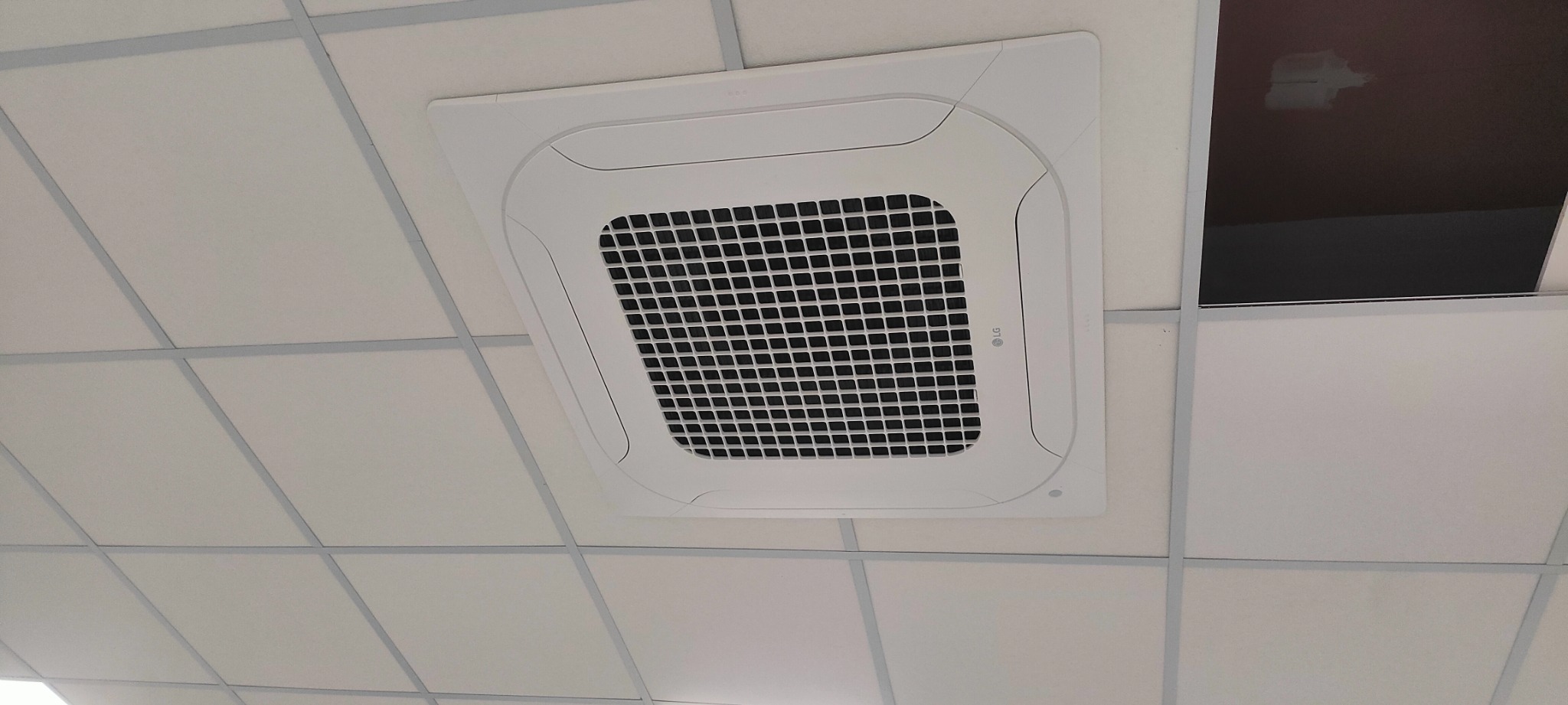 Kasetonowa klimatyzacja sufitowa LG w biurze, widok z dołu. Biała obudowa urządzenia wkomponowana w biały sufit podwieszany. Część sufitu jest uszkodzona.