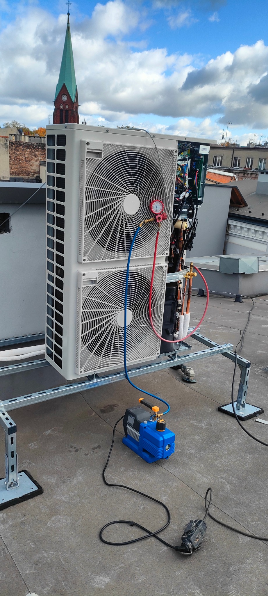 Dwuczęściowa jednostka zewnętrzna klimatyzacji na dachu, podłączona do pompy próżniowej. W tle wieża kościoła i zabudowa miejska.