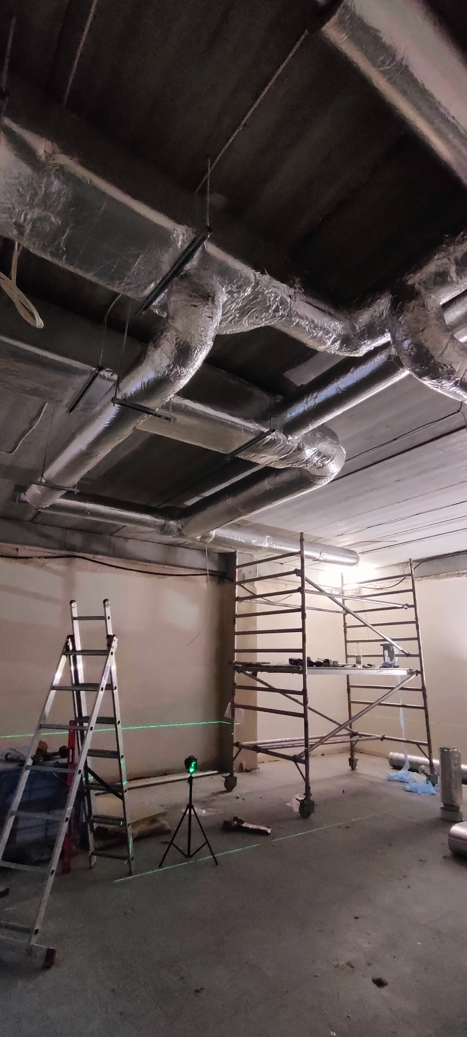 Montaż instalacji wentylacyjnej z użyciem rusztowania i poziomicy laserowej. Widoczne srebrne rury wentylacyjne pod sufitem i drabina oparta o ścianę.