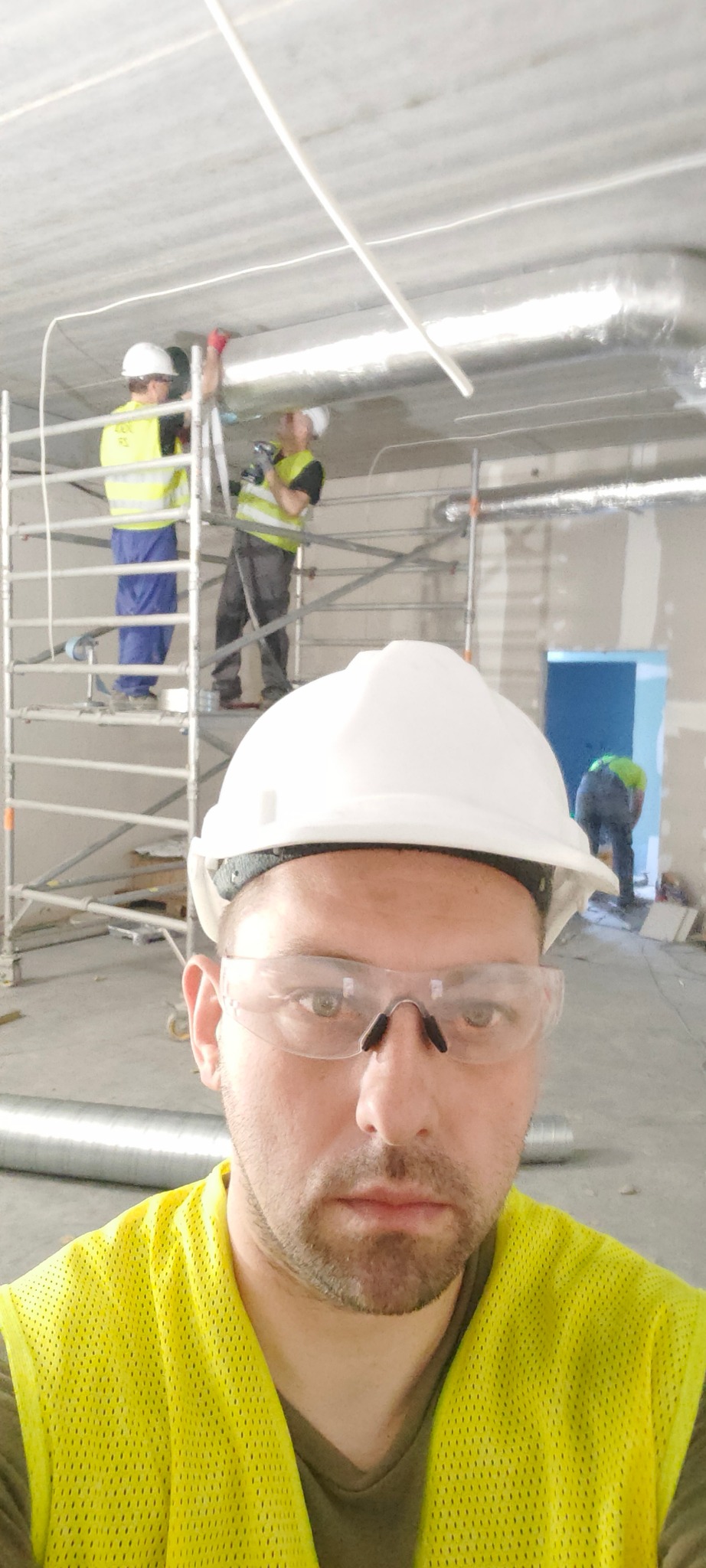 Instalacja wentylacji w budynku komercyjnym. Pracownicy na rusztowaniu montują srebrne rury wentylacyjne pod sufitem. Jeden z pracowników robi selfie.