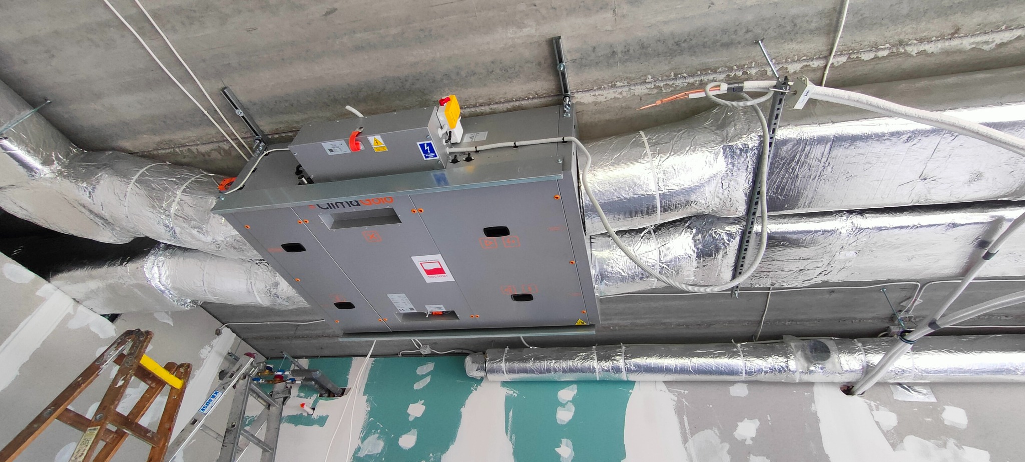 Szara centrala wentylacyjna Clima Gold zamontowana pod sufitem, z podłączonymi przewodami wentylacyjnymi w srebrnej izolacji. Widoczna drabina i ślady prac wykończeniowych.