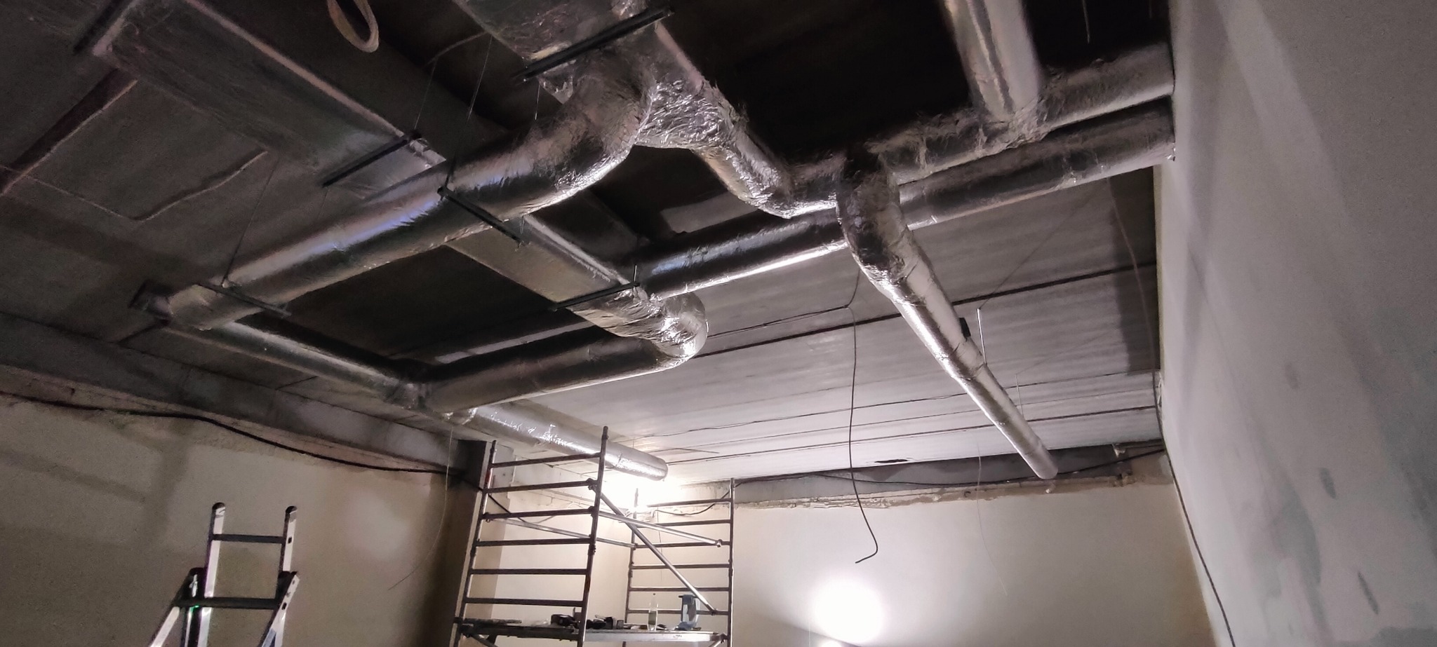 Srebrne, zaizolowane rury wentylacyjne pod sufitem w surowym wnętrzu. Widoczny fragment rusztowania i drabiny.