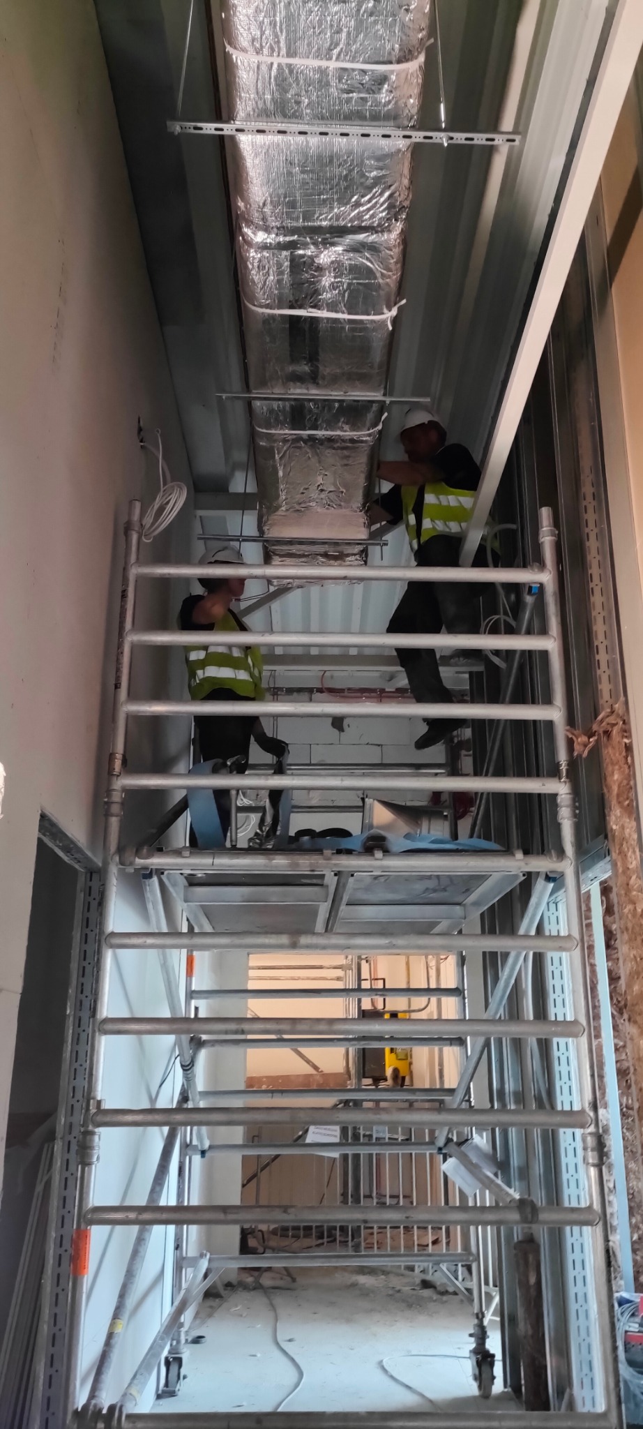 Montaż srebrnych przewodów wentylacyjnych przez dwóch monterów na rusztowaniu w surowym wnętrzu budynku. Widoczne elementy instalacji i narzędzia.