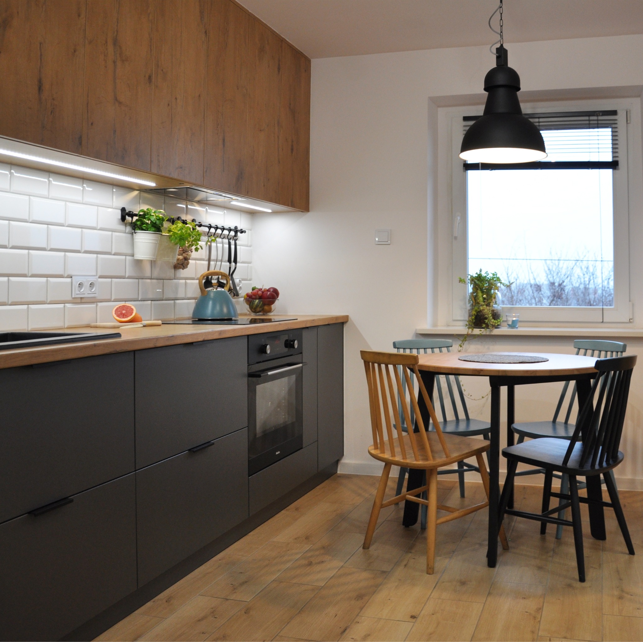 Nowoczesna kuchnia z ciemnoszarymi szafkami, drewnianym blatem, białymi płytkami metra i okrągłym stołem z różnokolorowymi krzesłami.