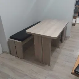 Stół z blatem imitującym jasne drewno, z dopasowaną ławką z czarnym siedziskiem, ustawiony na panelach podłogowych w odcieniach beżu i szarości.