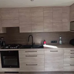 Jasna kuchnia z drewnopodobnymi frontami, czarną armaturą i piekarnikiem, widoczny kalendarz na ścianie.