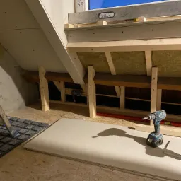 Montaż izolacji poddasza z widoczną konstrukcją drewnianą, wełną mineralną, płytą gipsowo-kartonową i wkrętarką akumulatorową.