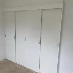 Biała szafa wnękowa z przesuwnymi drzwiami, widoczna na tle surowej podłogi z desek, wnętrze w trakcie wykończenia.