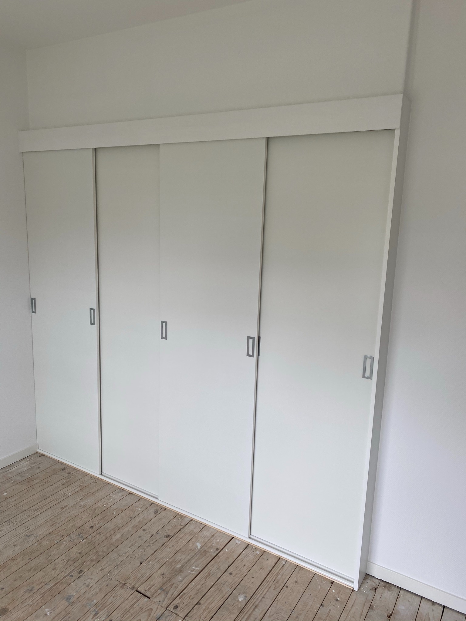 Biała szafa wnękowa z przesuwnymi drzwiami, widoczna na tle surowej podłogi z desek, wnętrze w trakcie wykończenia.