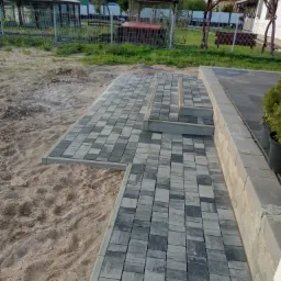 Ułożona ścieżka z szaro-czarnej kostki brukowej prowadząca do schodów, z widocznym przygotowanym podłożem z piasku i zielenią w tle.