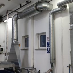 IMES Instalacje Sp. z o.o. - System wentylacji przemysłowej z metalowymi rurami i okapem, widoczne znaki BHP na ścianie, hala produkcyjna z bramą rolowaną w tle.