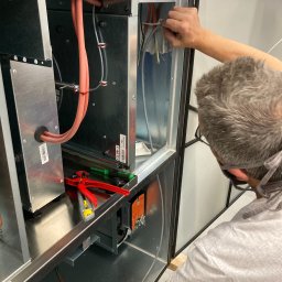 IMES Instalacje Sp. z o.o. - Instalator w okularach ochronnych podłącza przewody wewnątrz metalowej obudowy urządzenia wentylacyjnego w hali przemysłowej, widoczne narzędzia i kable.