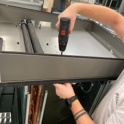 IMES Instalacje Sp. z o.o. - Montaż elementu instalacji wentylacyjnej: pracownik w białej koszulce przykręca śrubokrętem element obudowy rekuperatora, widoczne miedziane rurki wymiennika ciepła i czarne rury izolowane.