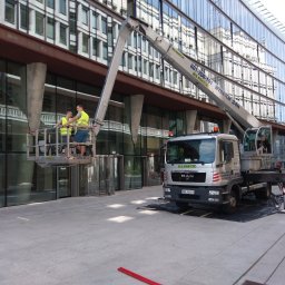 IMES Instalacje Sp. z o.o. - Dwóch pracowników w żółtych kamizelkach na podnośniku koszowym firmy Klimex czyści szklaną fasadę nowoczesnego biurowca, widoczny pojazd podnośnika z numerem rejestracyjnym WW 1669H.