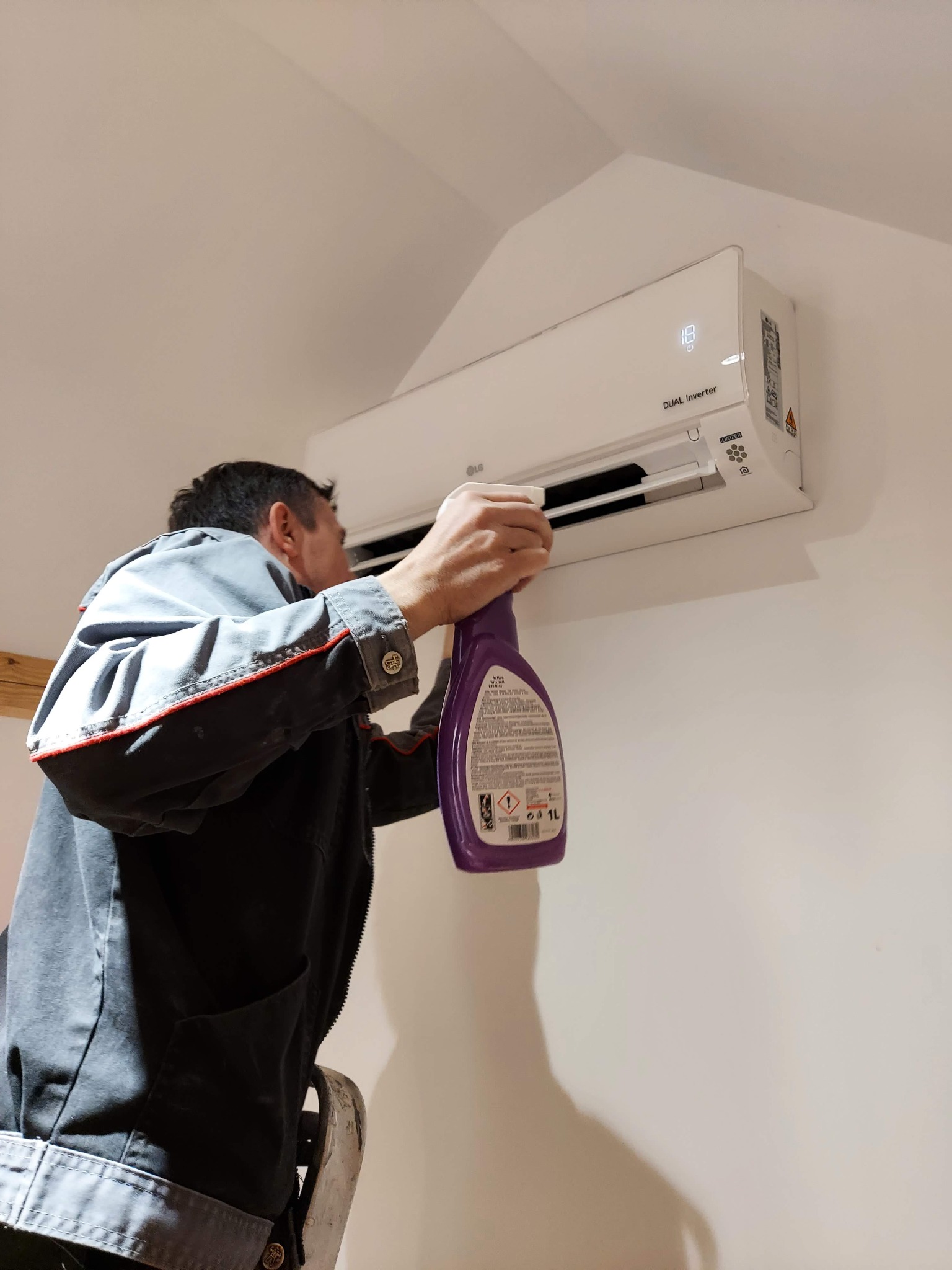 Mężczyzna w szarej kurtce czyści klimatyzator LG Dual Inverter za pomocą fioletowego sprayu czyszczącego.