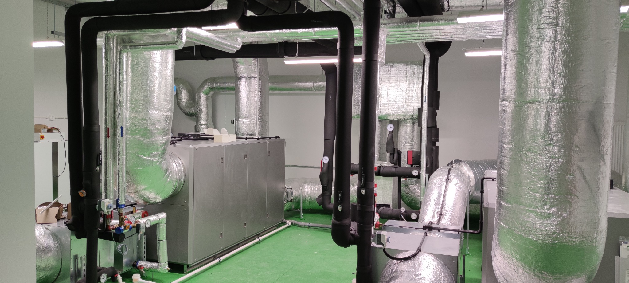 Centrala wentylacyjna z izolowanymi przewodami powietrznymi i rurami w pomieszczeniu technicznym, widoczne manometry na instalacji.