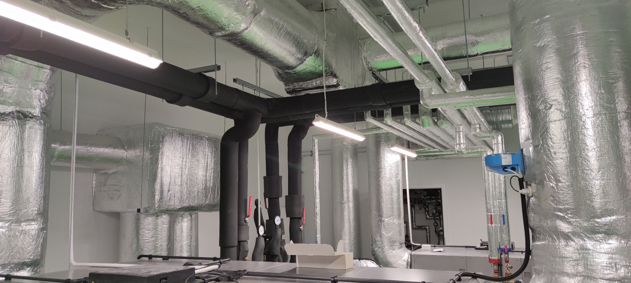 Instalacja rur wentylacyjnych i hydraulicznych w budynku komercyjnym, z izolacją termiczną, widoczne zawory i manometry.