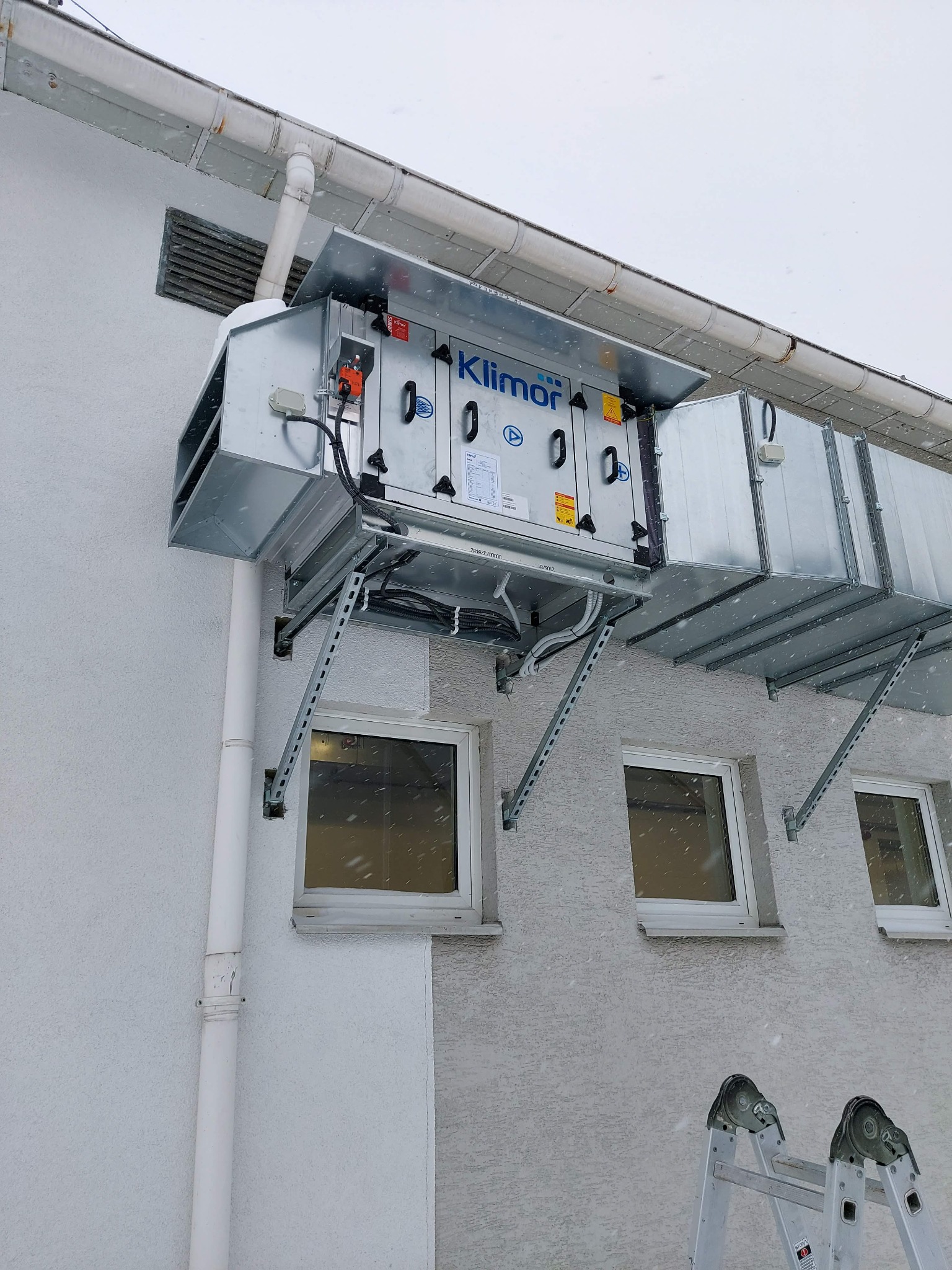Zewnętrzna instalacja rekuperacji marki Klimor na ścianie budynku w zimowej scenerii, widoczna drabina sugerująca prace montażowe lub serwisowe.