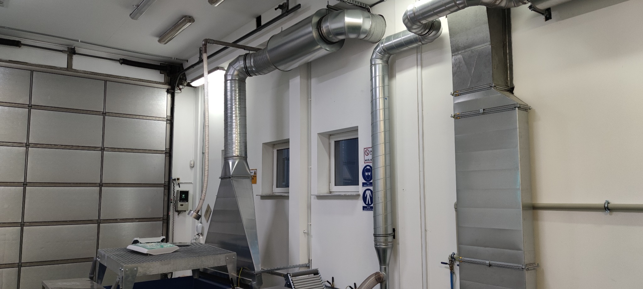 System wentylacji przemysłowej z metalowymi rurami i okapem, widoczne znaki BHP na ścianie, hala produkcyjna z bramą rolowaną w tle.
