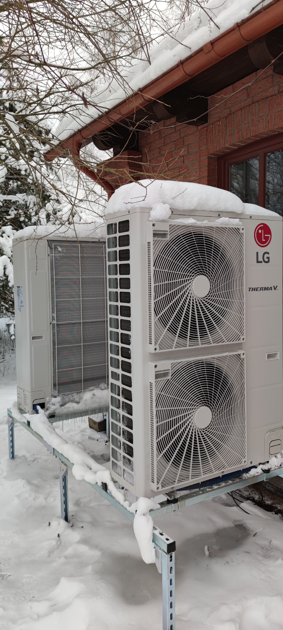 Dwa białe moduły pompy ciepła LG Therma V, częściowo zasypane śniegiem, stojące na metalowej konstrukcji na zewnątrz budynku z cegły w zimowej scenerii.
