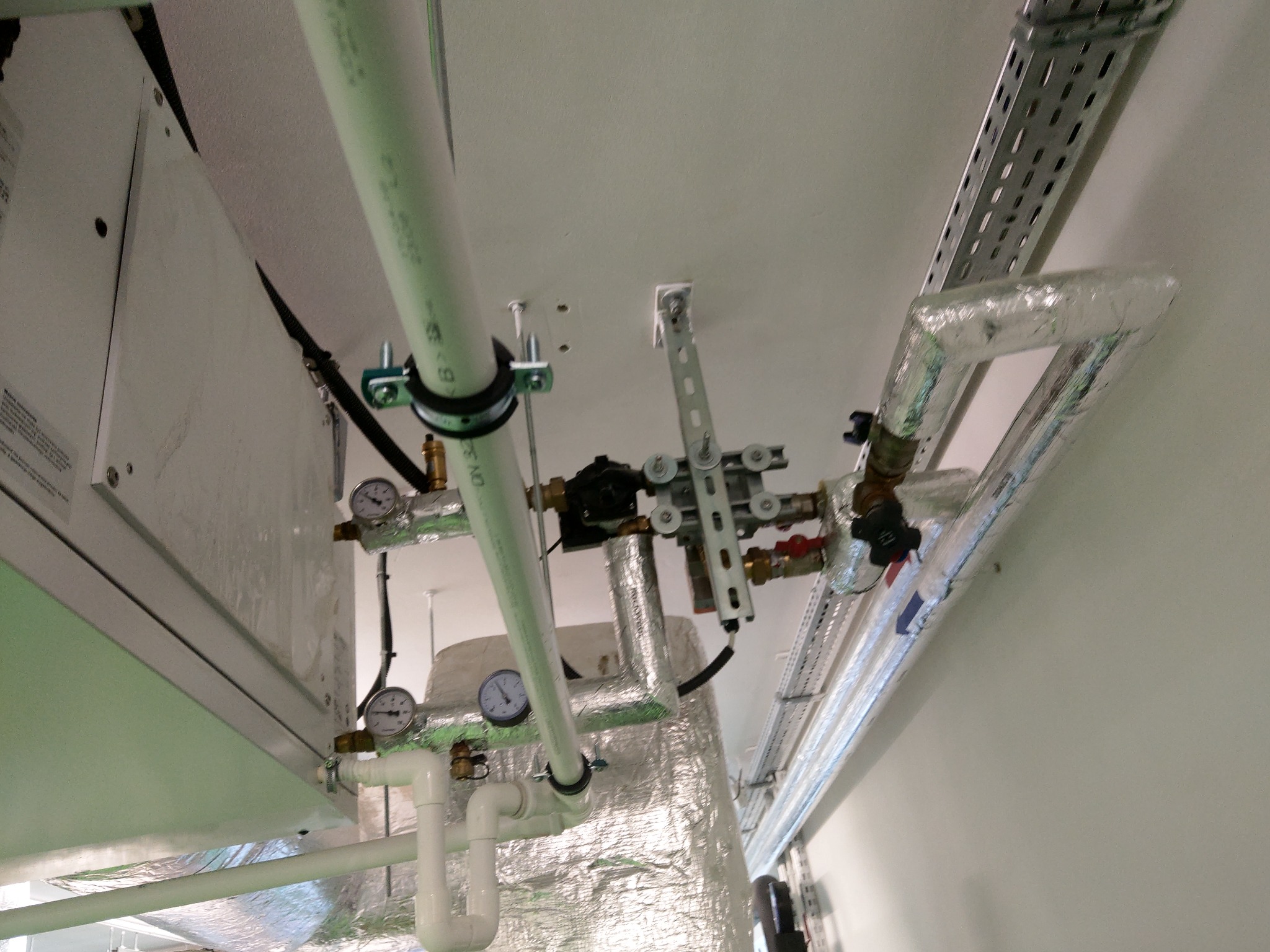 Instalacja hydrauliczna pod sufitem z rurami, zaworami, manometrami i izolacją termiczną, widoczne elementy mocujące i korytka kablowe.