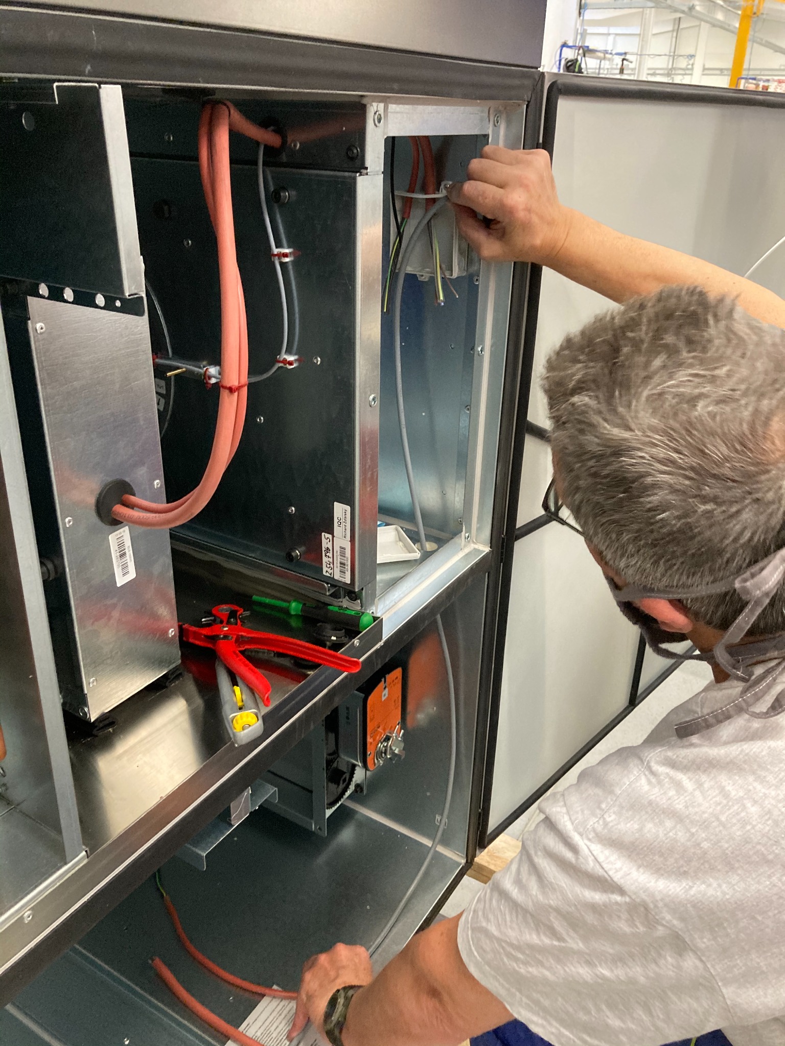 Instalator w okularach ochronnych podłącza przewody wewnątrz metalowej obudowy urządzenia wentylacyjnego w hali przemysłowej, widoczne narzędzia i kable.