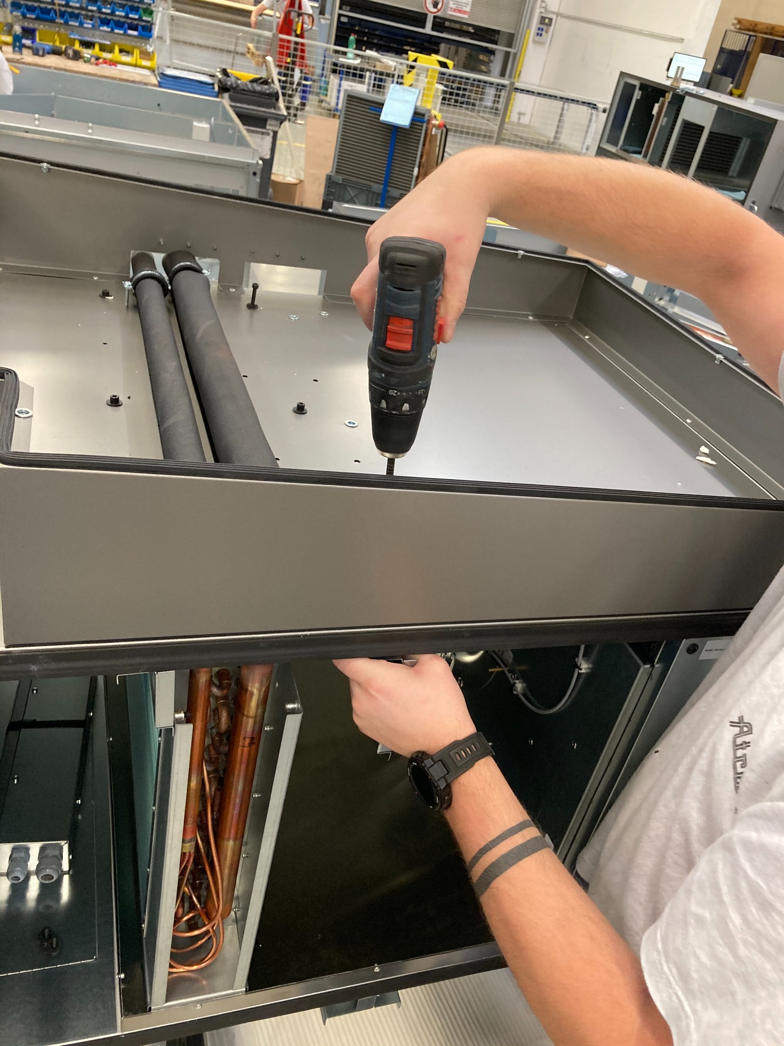 Montaż elementu instalacji wentylacyjnej: pracownik w białej koszulce przykręca śrubokrętem element obudowy rekuperatora, widoczne miedziane rurki wymiennika ciepła i czarne rury izolowane.