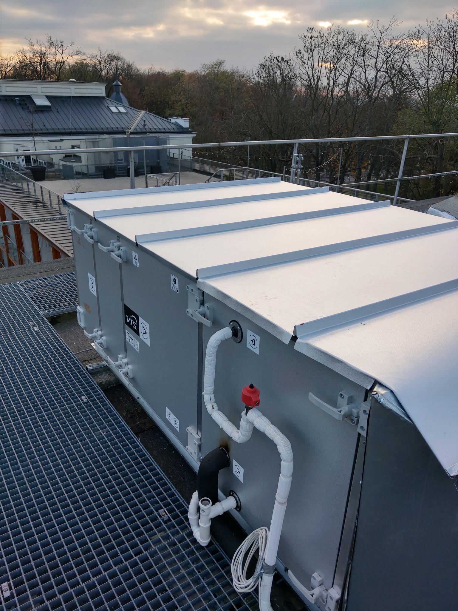 Centrala wentylacyjna z rurami na dachu budynku, widok z góry, w tle drzewa i budynki, pochmurne niebo.