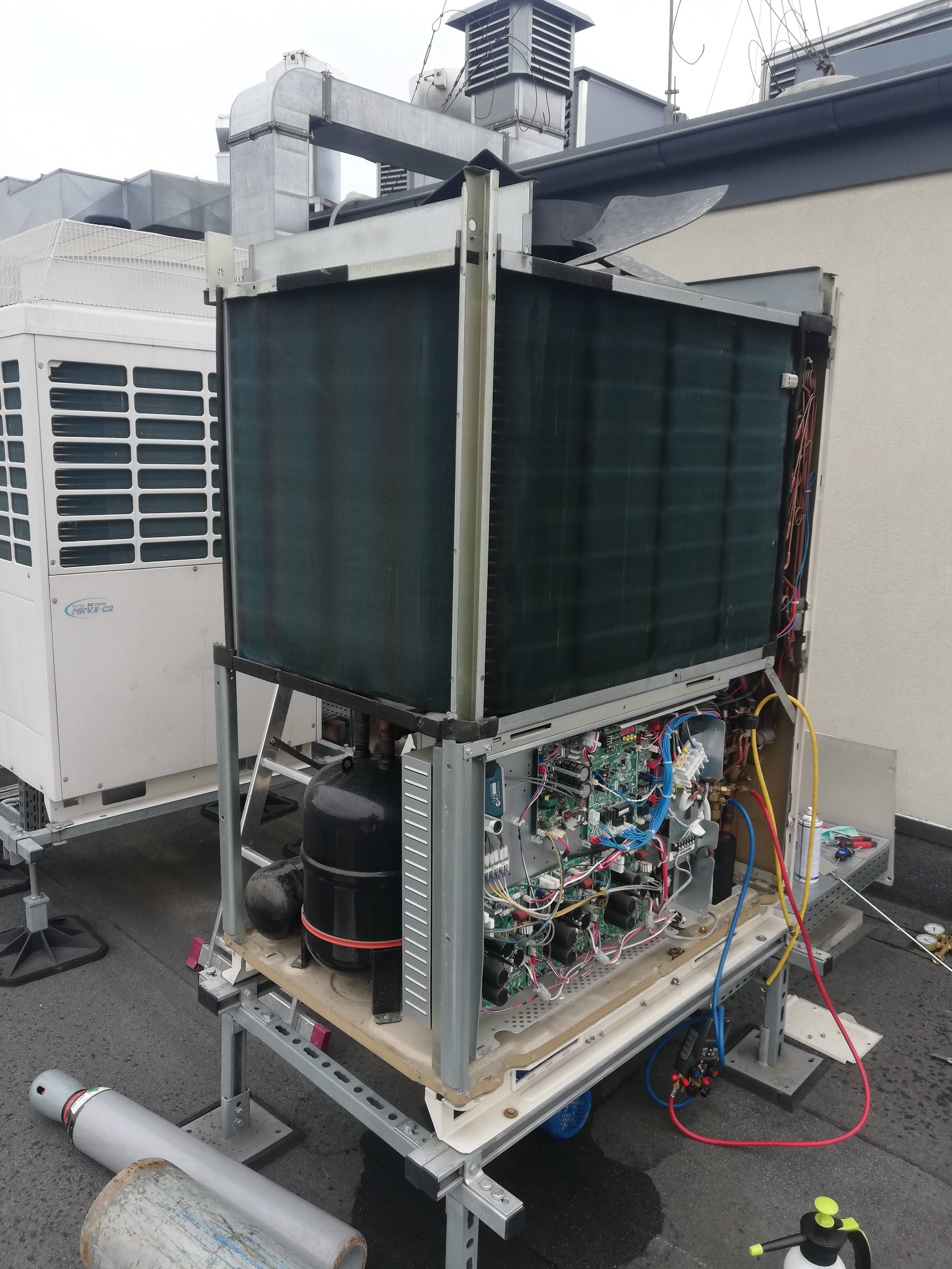 Otwarta jednostka klimatyzacyjna na dachu budynku, widoczne wewnętrzne komponenty, w tym elektronika i rury, obok butle z gazem i narzędzia.