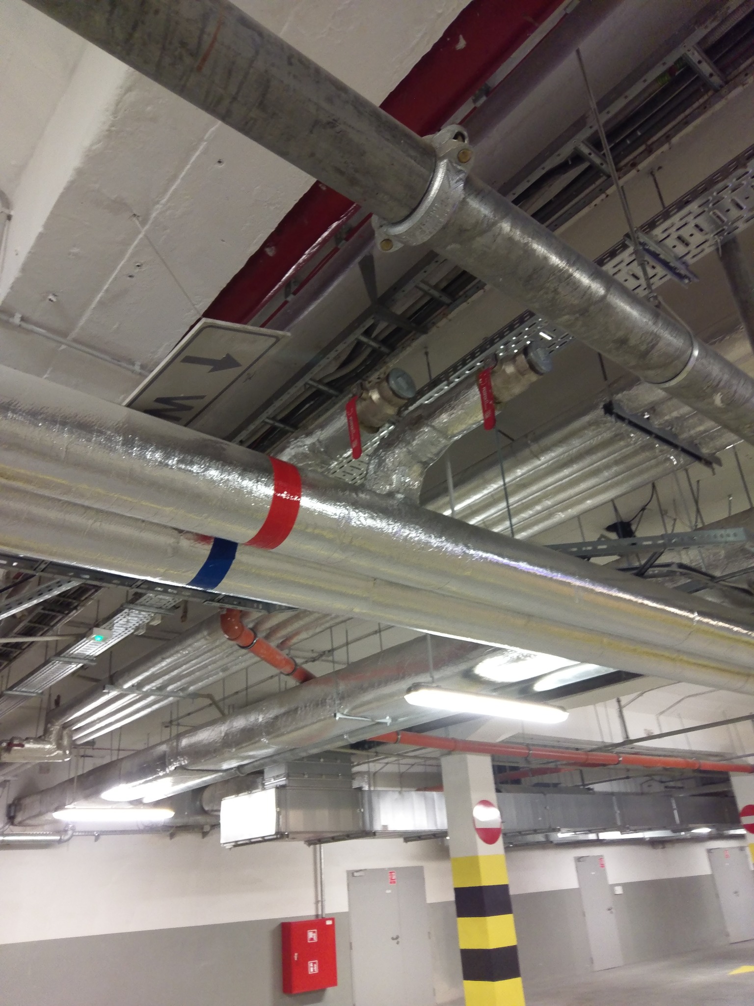 Instalacje wentylacyjne i hydrauliczne pod sufitem parkingu podziemnego, rury z izolacją aluminiową, zawory z czerwonymi uchwytami, kable w korytach, oznaczenia kolorystyczne na rurach.