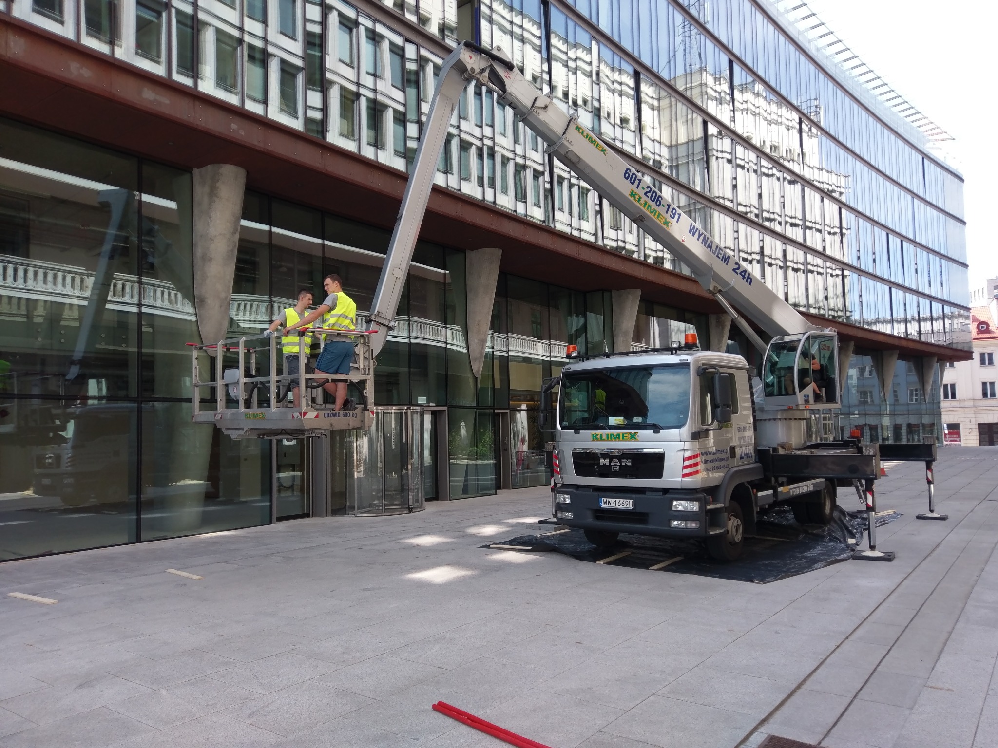 Dwóch pracowników w żółtych kamizelkach na podnośniku koszowym firmy Klimex czyści szklaną fasadę nowoczesnego biurowca, widoczny pojazd podnośnika z numerem rejestracyjnym WW 1669H.