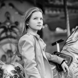 Justyna Tomczak Photography - Czarno-białe zdjęcie nastolatki w jasnej kurtce, siedzącej na karuzelowym koniu z ręcznie malowanymi kwiatami, zamyślona poza i wiatr we włosach.