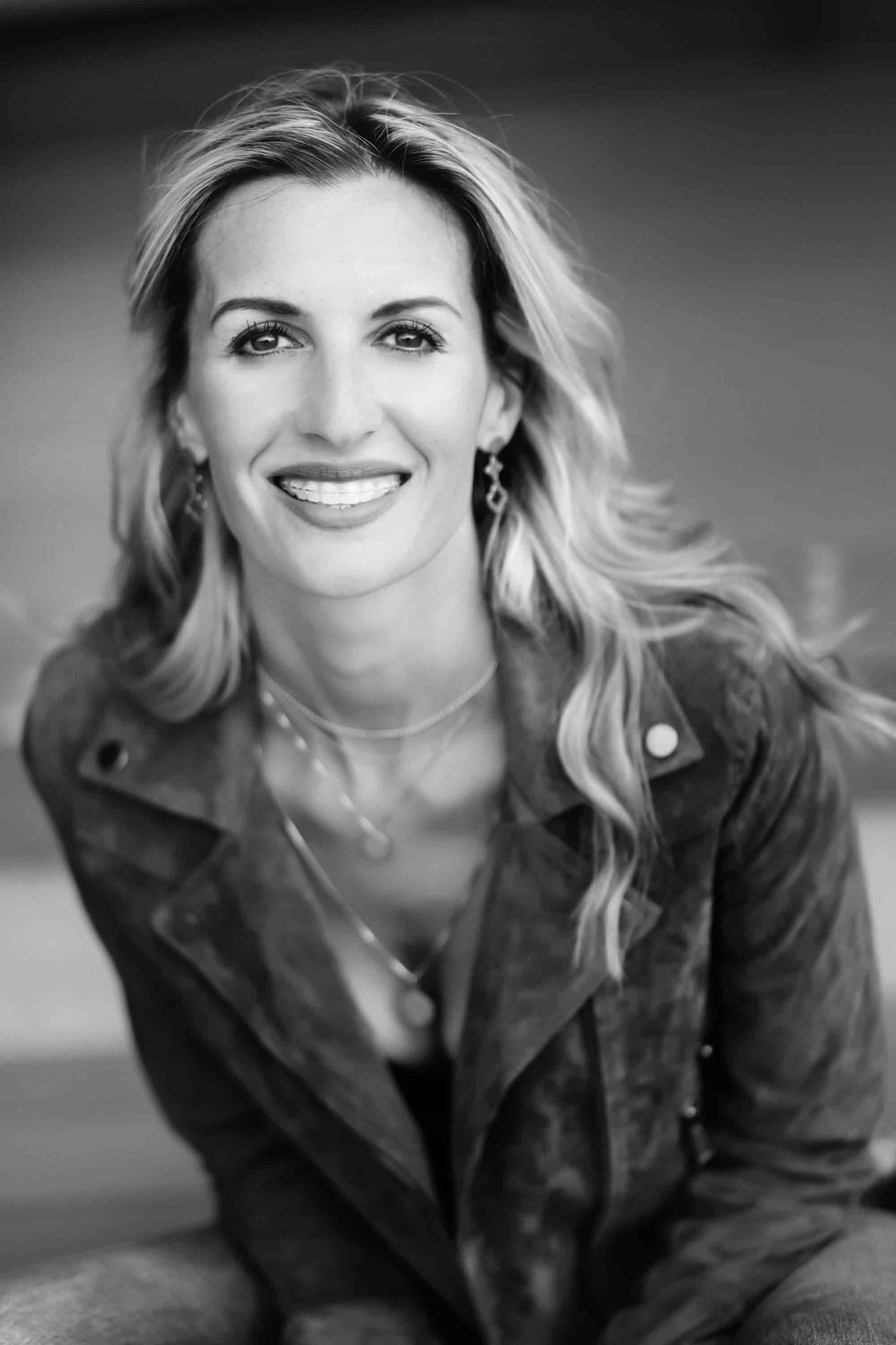 Portret uśmiechniętej kobiety w czarno-białej tonacji, z widocznym aparatem ortodontycznym, noszącej skórzaną kurtkę i delikatną biżuterię, z rozpuszczonymi, falowanymi włosami.