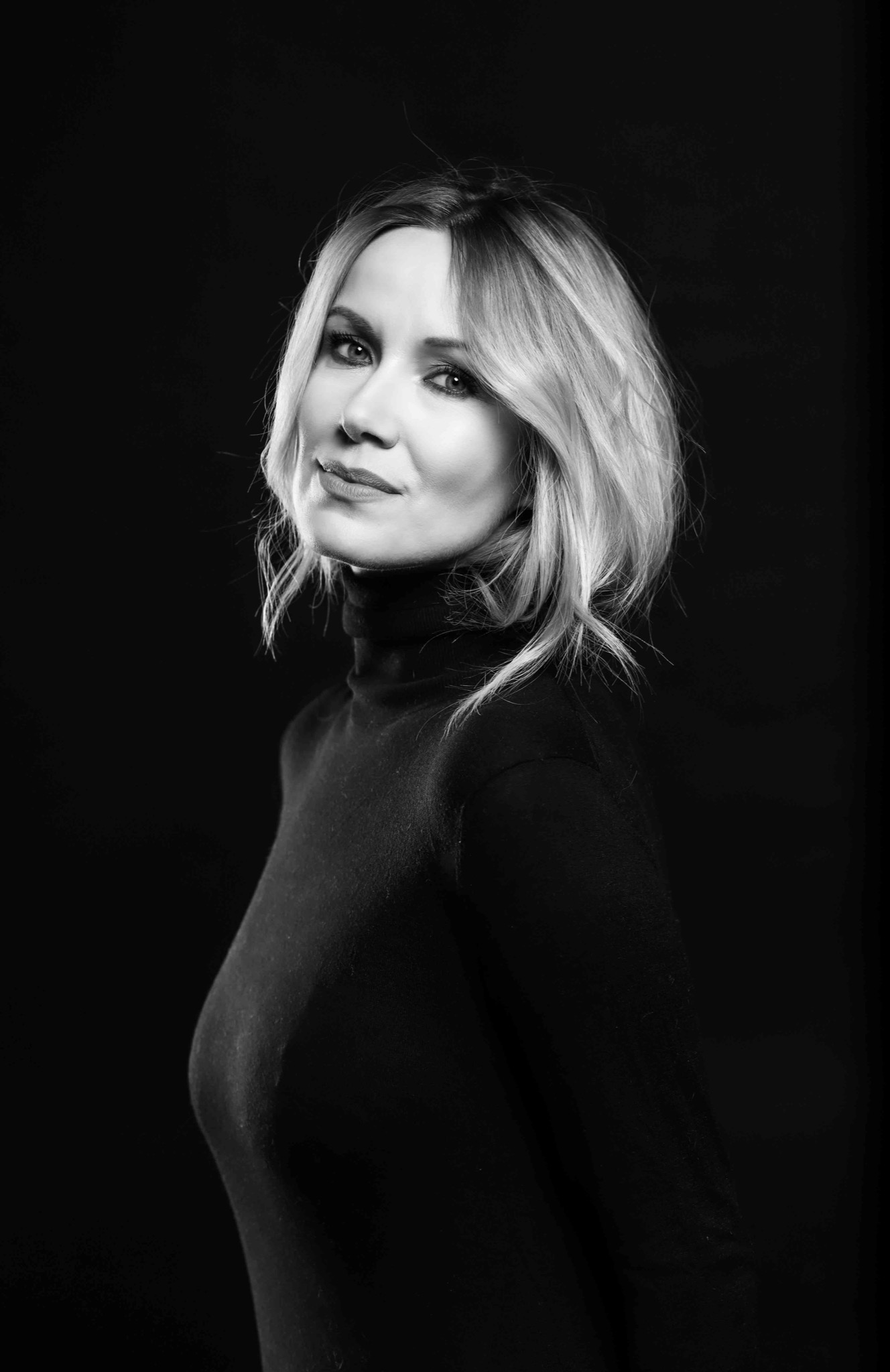 Portret kobiety z blond włosami w czarnym golfie na ciemnym tle, uchwycony w czerni i bieli, z delikatnym uśmiechem i spojrzeniem skierowanym w górę.